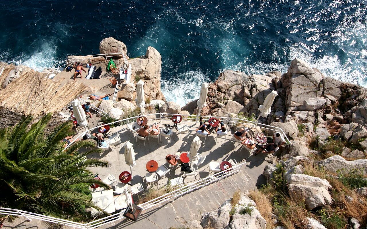 image source Photo by Martin Uradnicek: https://www.pexels.com/photo/tourists-on-the-terraces-of-the-buza-bar-on-the-seaside-cliff-of-dubrovnik-12383597/